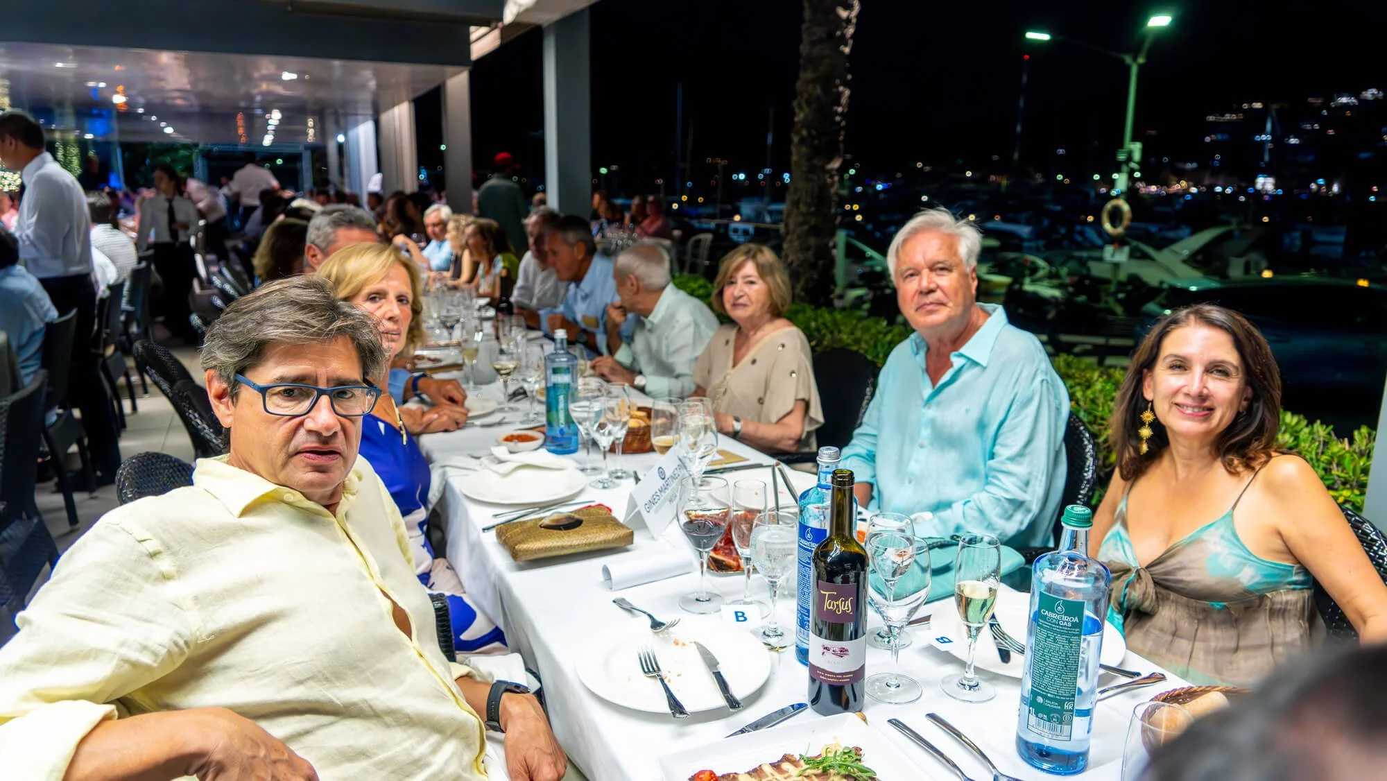 Un grupo de personas sentadas en una mesa de restaurante durante la noche, con comida y bebidas frente a ellos, en un ambiente elegante y social.