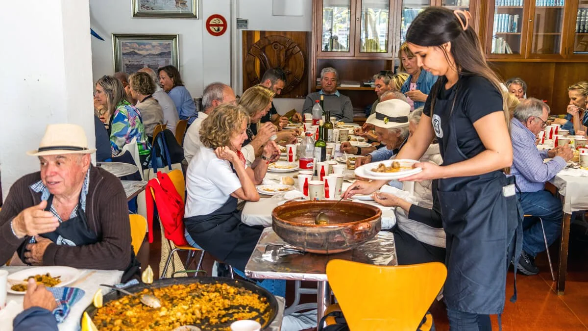 Mesa larga con muchas personas comiendo en un comedor, una joven sirviendo comida desde un recipiente grande.