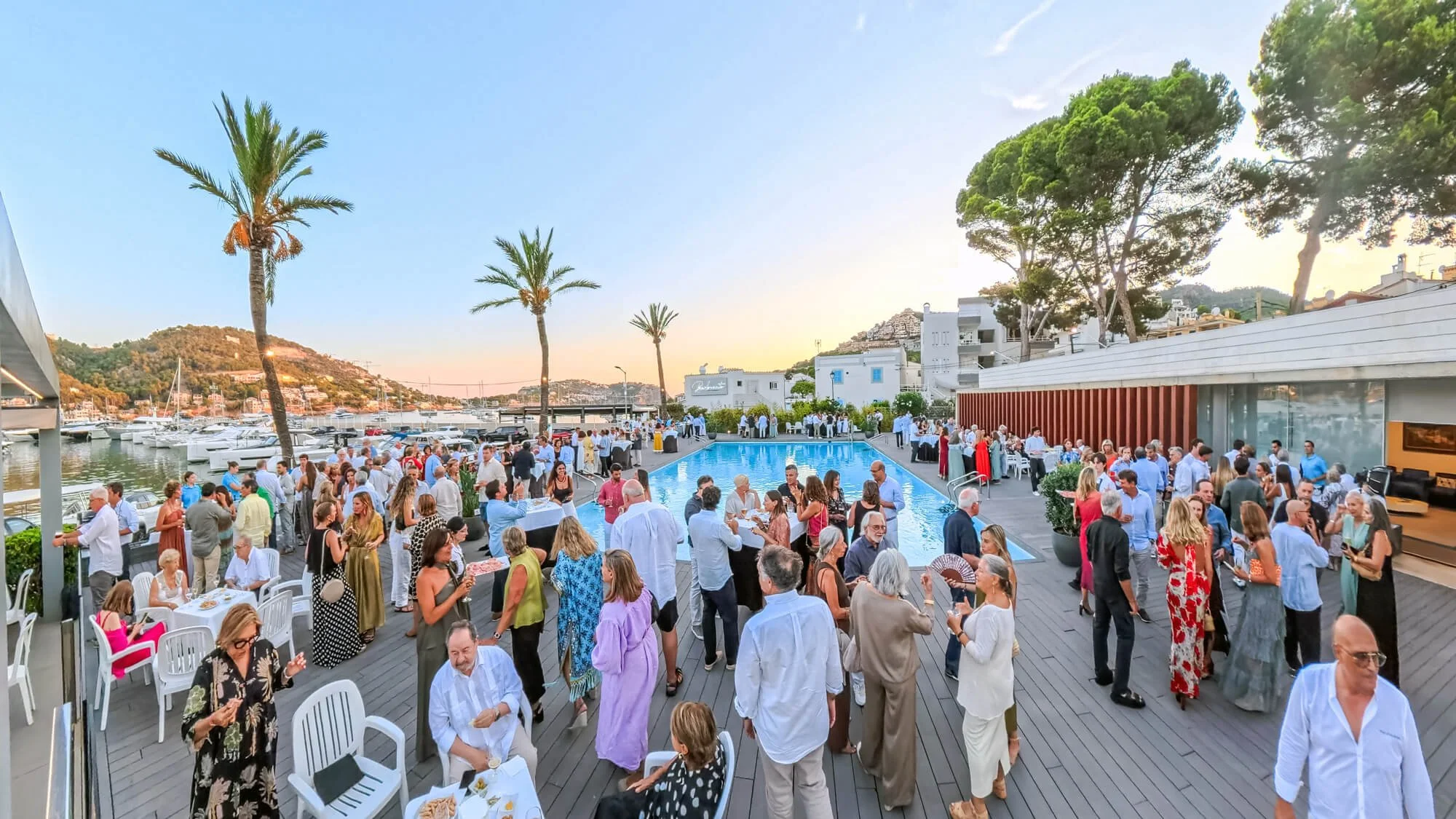 Cena de una fiesta al aire libre junto a una piscina, con muchas personas vestidas de forma elegante, disfrutando y socializando, en un entorno con árboles, palmeras, montañas y yates en un puerto al fondo, durante el atardecer.