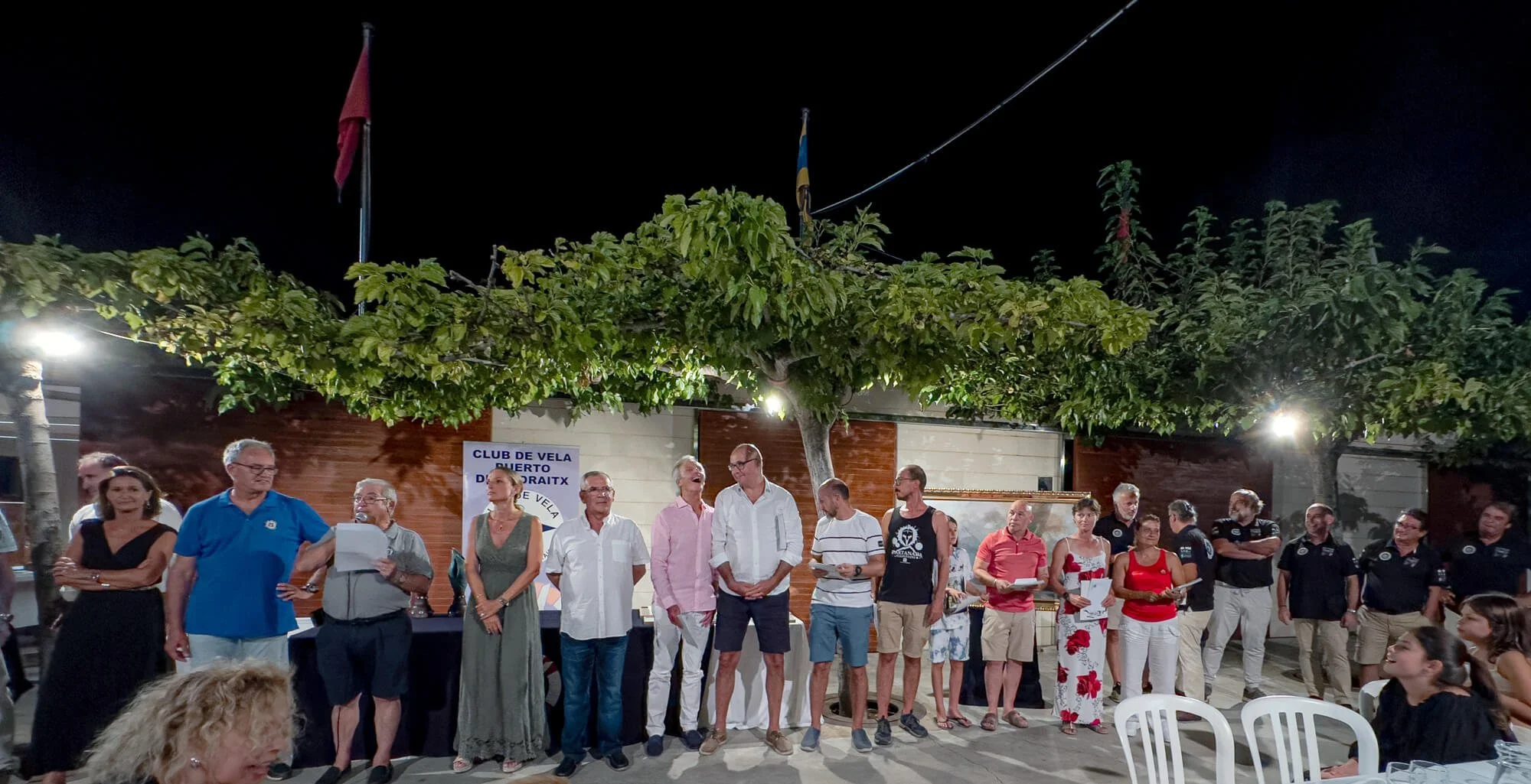 Personas reunidas en una ceremonia al aire libre en la noche, con un árbol en el fondo, en un evento del club de vela de Puerto Doroaitx.