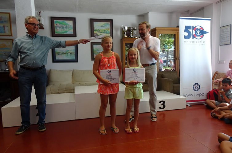 Dos niñas sosteniendo certificados en un escenario, con dos adultos entregando los premios, en un salón decorado con cuadros y un banner con el logo del 50 aniversario del CVPA. Niños observando en el suelo.