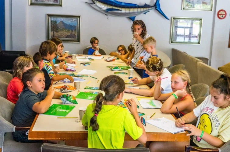 Niños en una mesa participando en una actividad artística en una sala decorada con cuadros y un pez enorme en la pared.