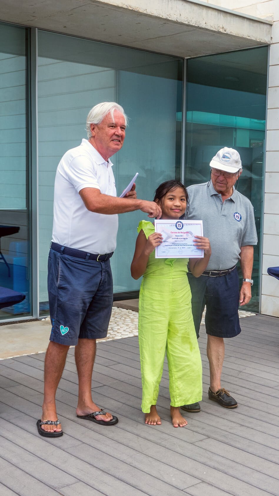 Una niña recibe un certificado de un curso de natación durante una ceremonia al aire libre, mientras dos adultos la acompañan.