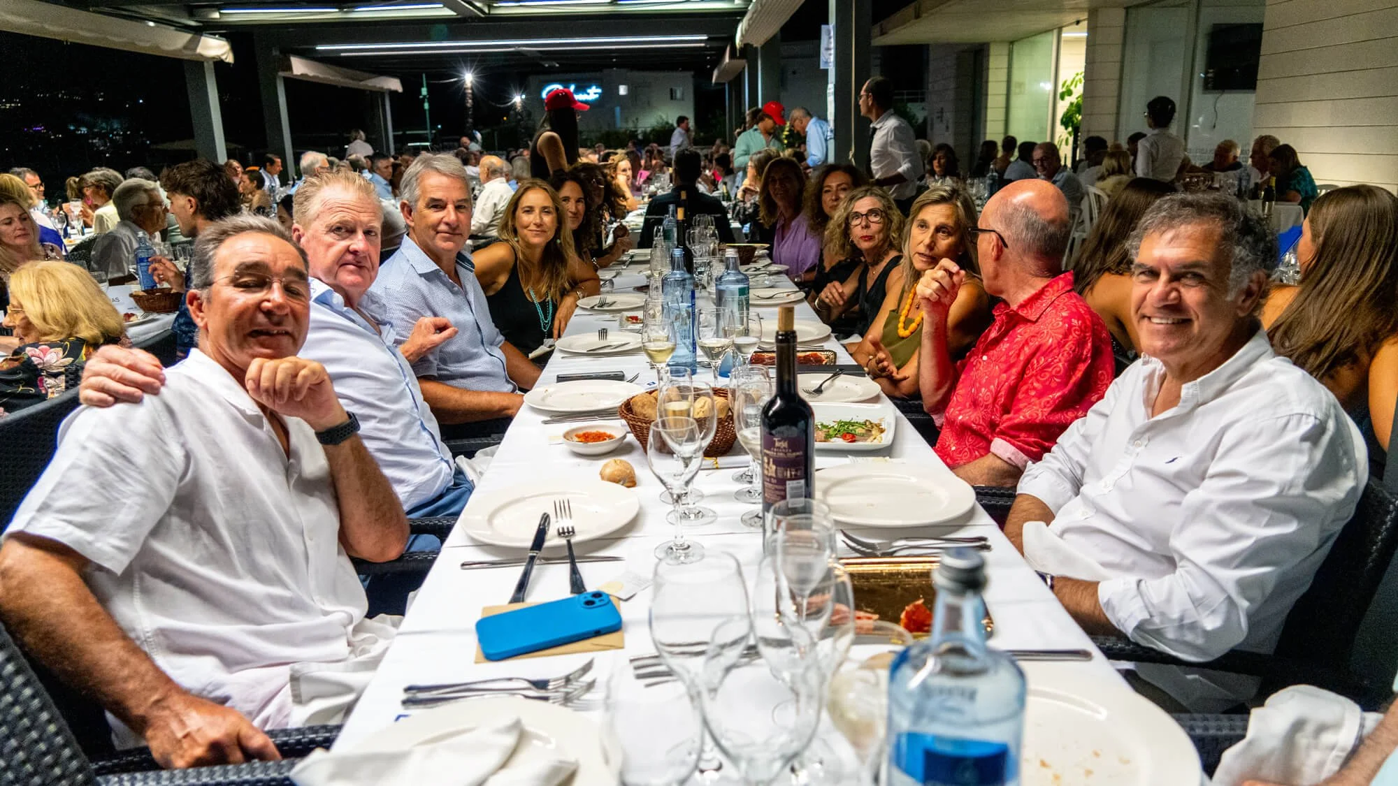 Grupo de personas sentadas en una mesa larga en un restaurante durante una celebración, con comida y bebidas en la mesa, algunos sonriendo y otros conversando, en un entorno interior nocturno.