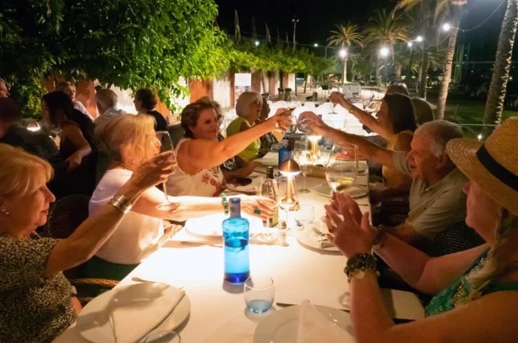 Grupo de personas mayores disfrutando una cena al aire libre en una noche con iluminación suave y palmeras en el fondo.