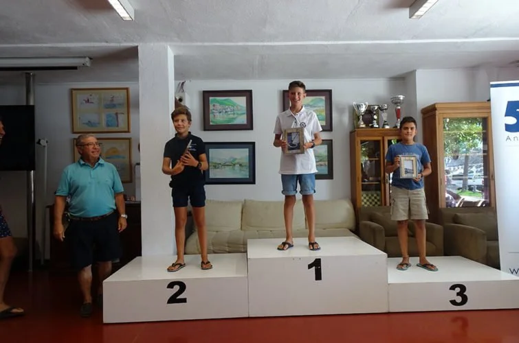 Niños en un podio de premios en una ceremonia, con los primeros, segundos y terceros lugares, en un ambiente interior con cuadros en la pared y muebles de madera.
