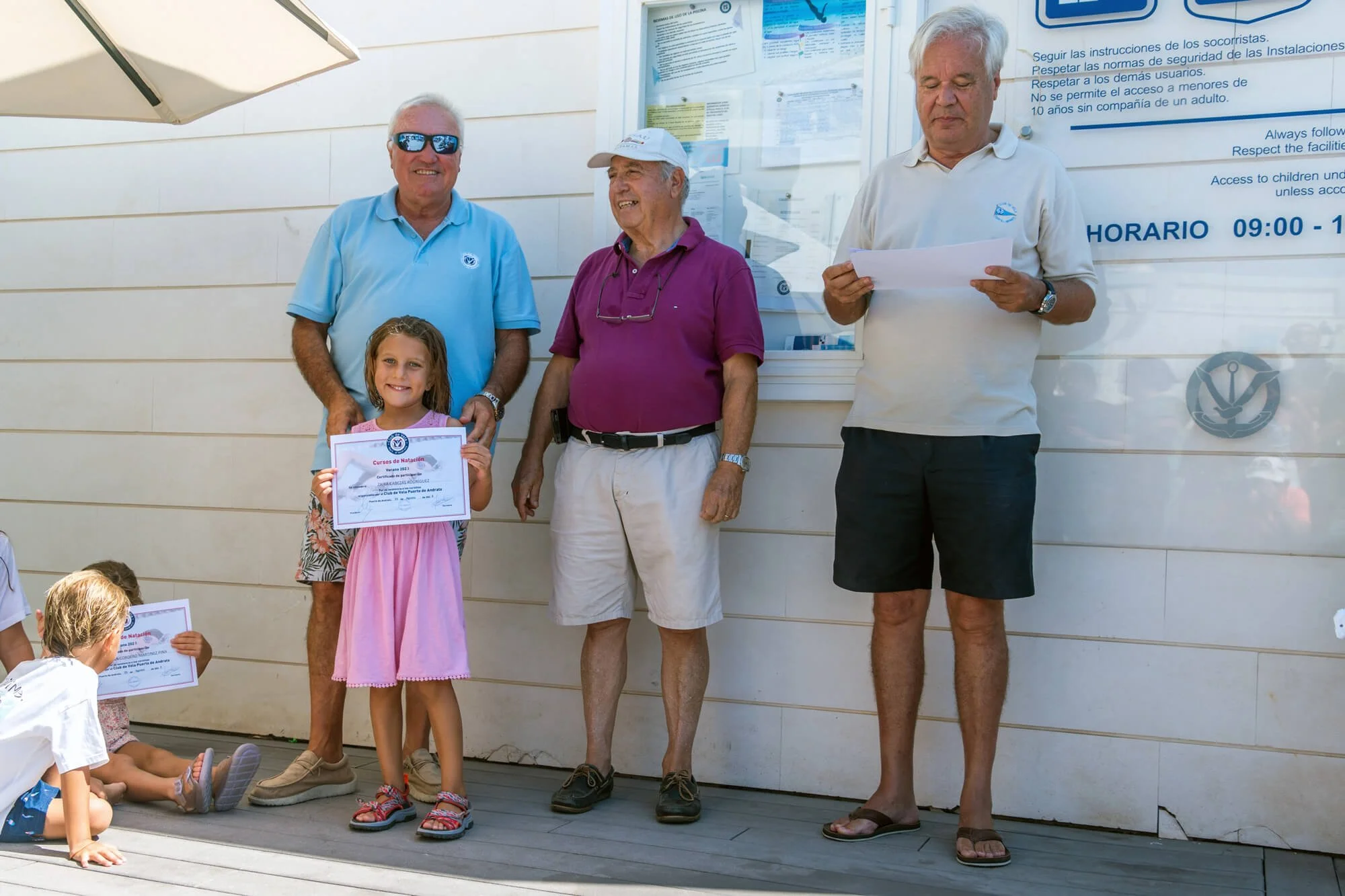 Niña recibiendo un certificado de natación en una ceremonia al aire libre, acompañada por tres adultos, uno con gafas de sol y otro con gafas y capa, frente a un cartel con instrucciones en español.