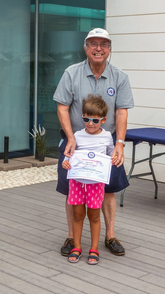 Un anciano y una niña posando juntos al aire libre. La niña sostiene un certificado y lleva gafas de sol, mientras que el anciano la sostiene por los hombros y ambos sonríen.