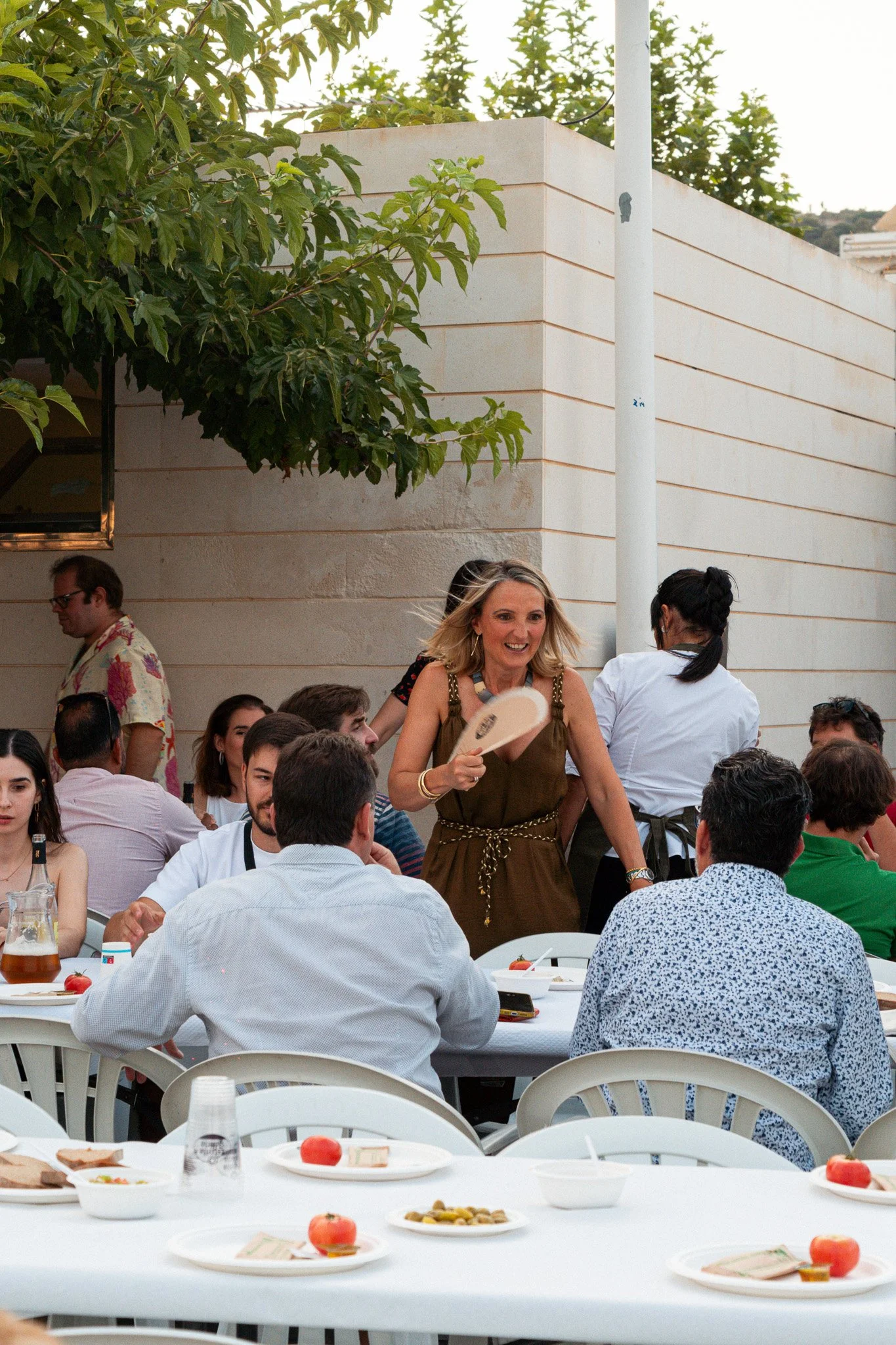 Una escena en un evento social al aire libre con muchas personas comiendo y sirviendo comida en una mesa grande, en el fondo una pared blanca y vegetación.