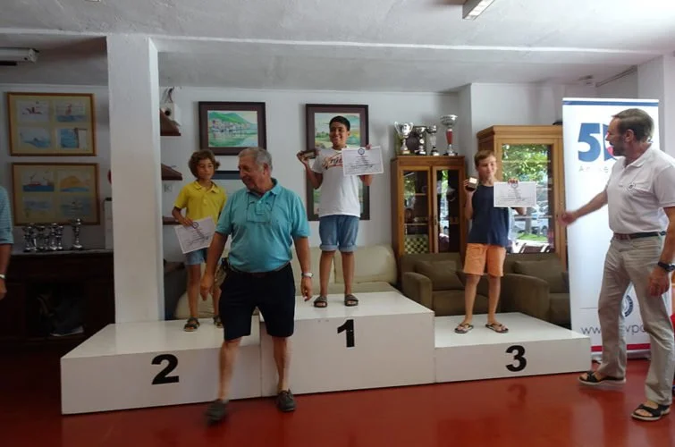 Niños en un podio de premios, con un trofeo y certificados, en una ceremonia de premiación, junto a adultos, en un interior con paredes blancas y cuadros en las paredes.