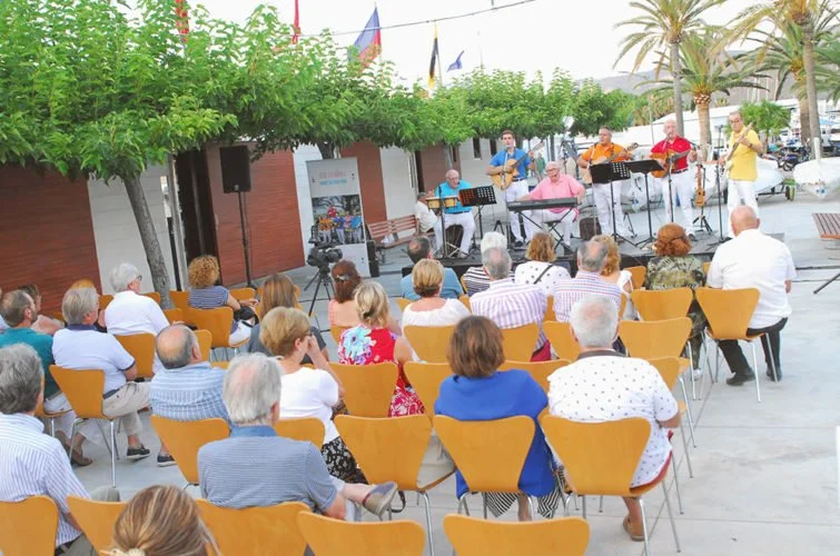 Concierto al aire libre con músicos tocando en un escenario y audiencia sentada disfrutando la música en una plaza con árboles y palmeras.