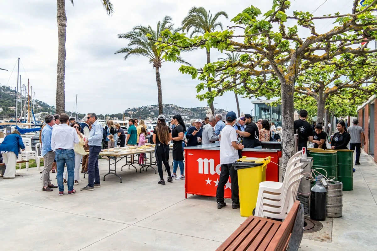 Gente disfrutando en una reunión al aire libre cerca del mar, con mesas, comida y bebidas, en un día nublado.