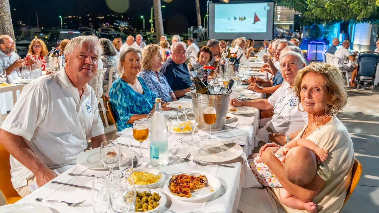 Personas sentadas en una mesa de cena al aire libre durante la noche, con comida y bebidas, en un evento social o celebración.