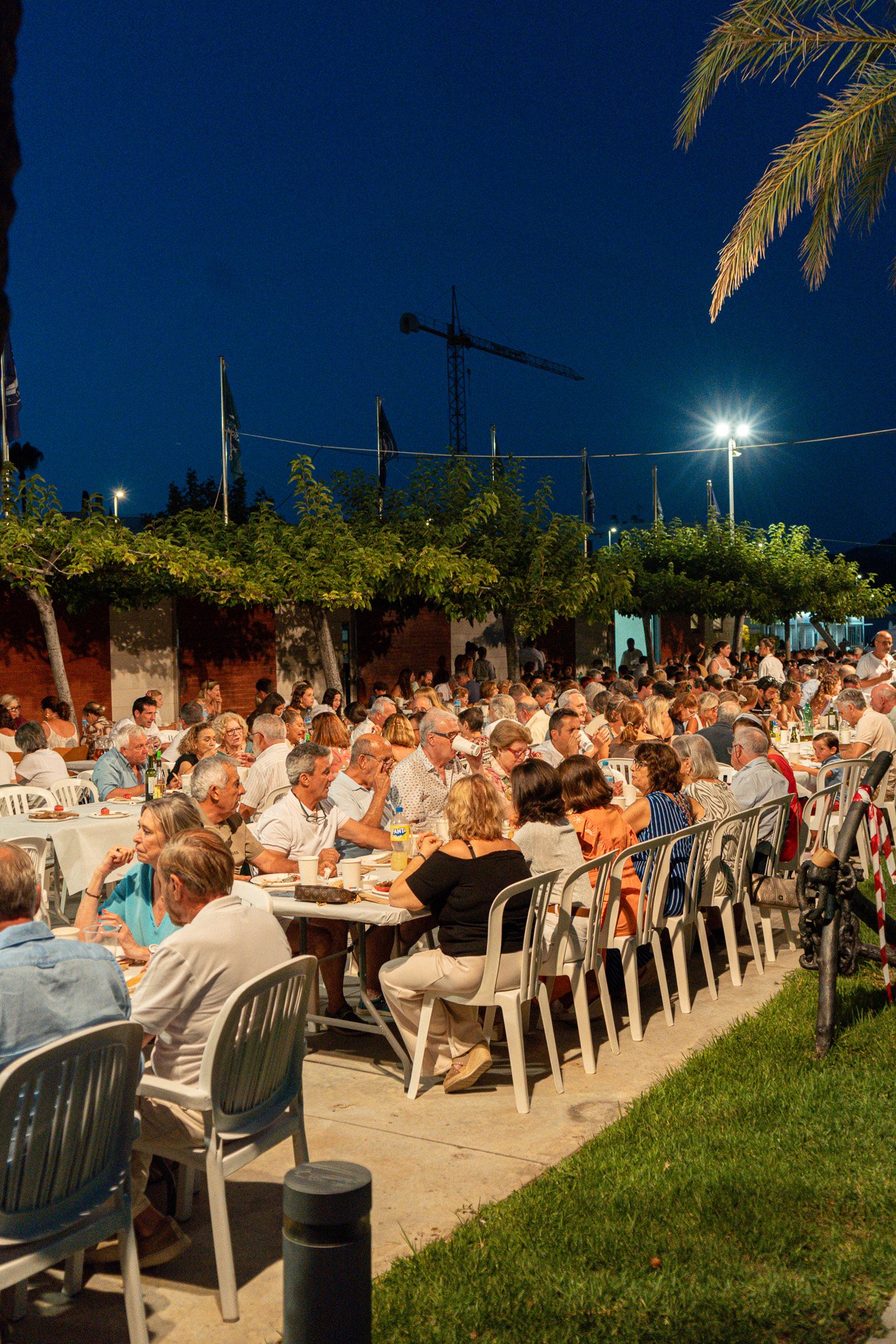 Gente cenando al aire libre en la noche, con árboles, luces y construcciones en el fondo.