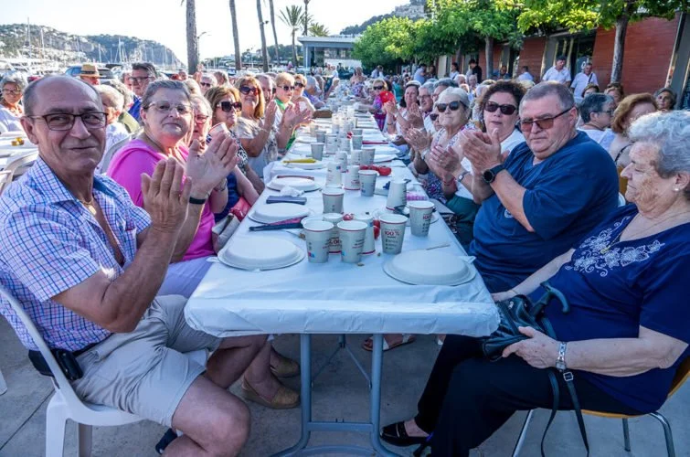 Grupo de personas mayores sentadas en una mesa al aire libre, disfrutando de una comida durante un evento social en la playa con palmeras y edificios de fondo.