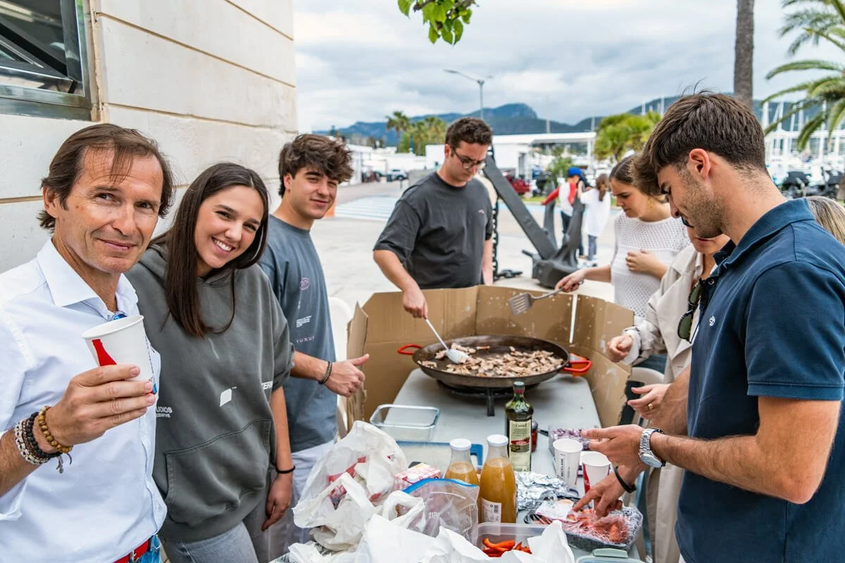 Grupo de jóvenes y un adulto en una reunión al aire libre, cocinando en una parrilla con carne y salsas, en un ambiente urbano con palmeras y montañas al fondo.