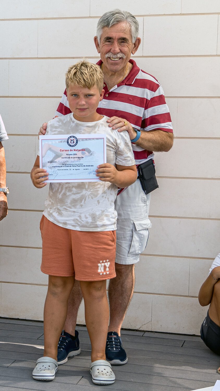 Un niño con camiseta blanca y pantalones cortos anaranjados sostiene un certificado, acompañado por un adulto mayor con camiseta de rayas rojas y blancas, en un evento de natación.