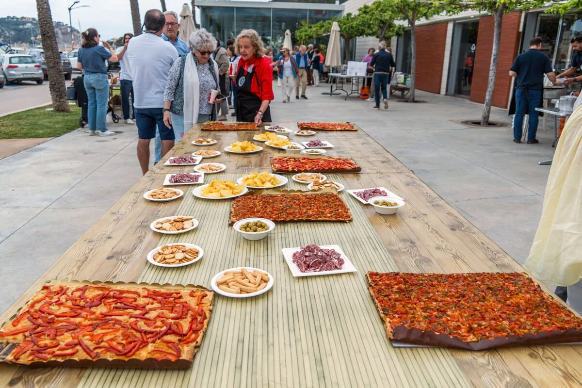 Mesa al aire libre con pizza y aperitivos, donde varias personas están disfrutando de una degustación en un evento.