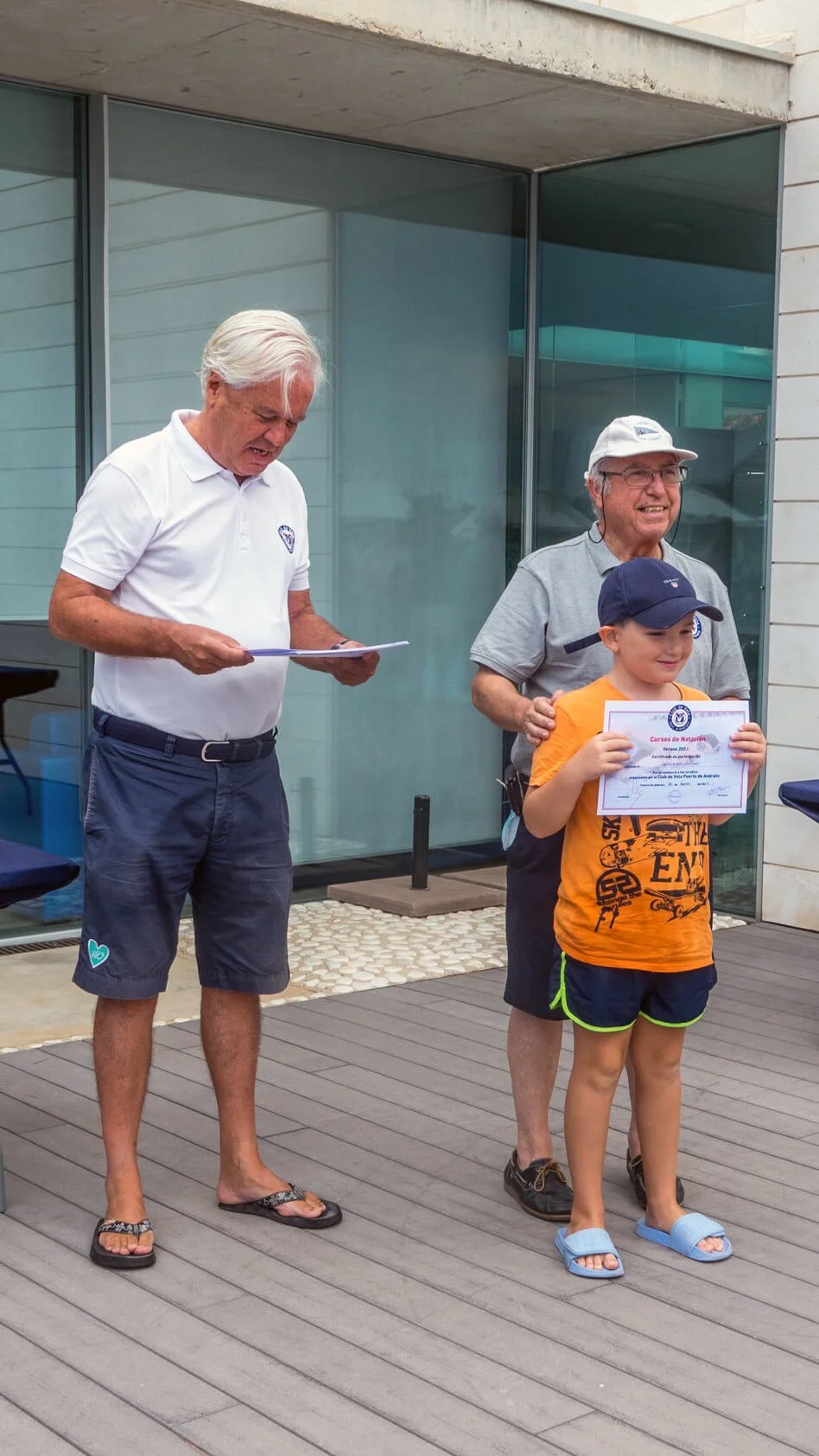 Un niño recibe un certificado de natación durante una ceremonia al aire libre, acompañado de dos adultos, uno de los cuales le está entregando el reconocimiento.