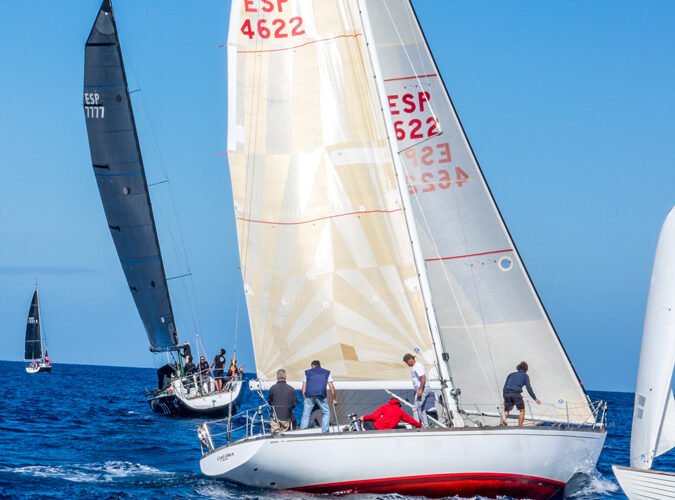 Vela de regata en el mar con varias personas a bordo, con muchas velas blancas y negras, y un cielo despejado.