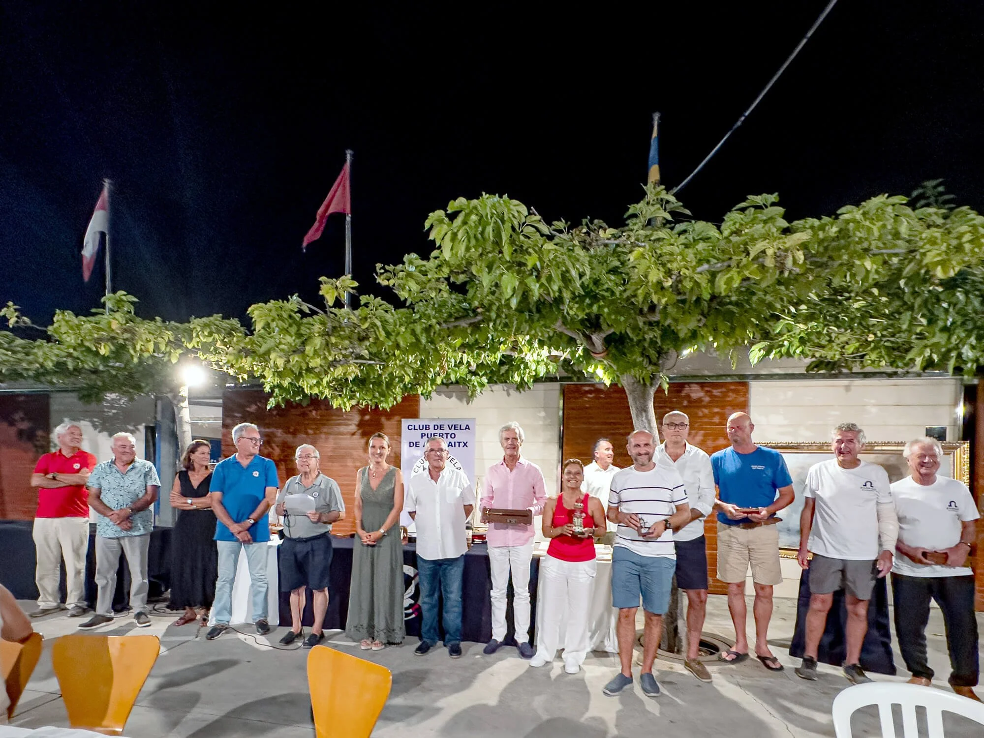 Grupo de personas en un evento al aire libre, con banderas en el fondo y un árbol decorativo, en la noche.