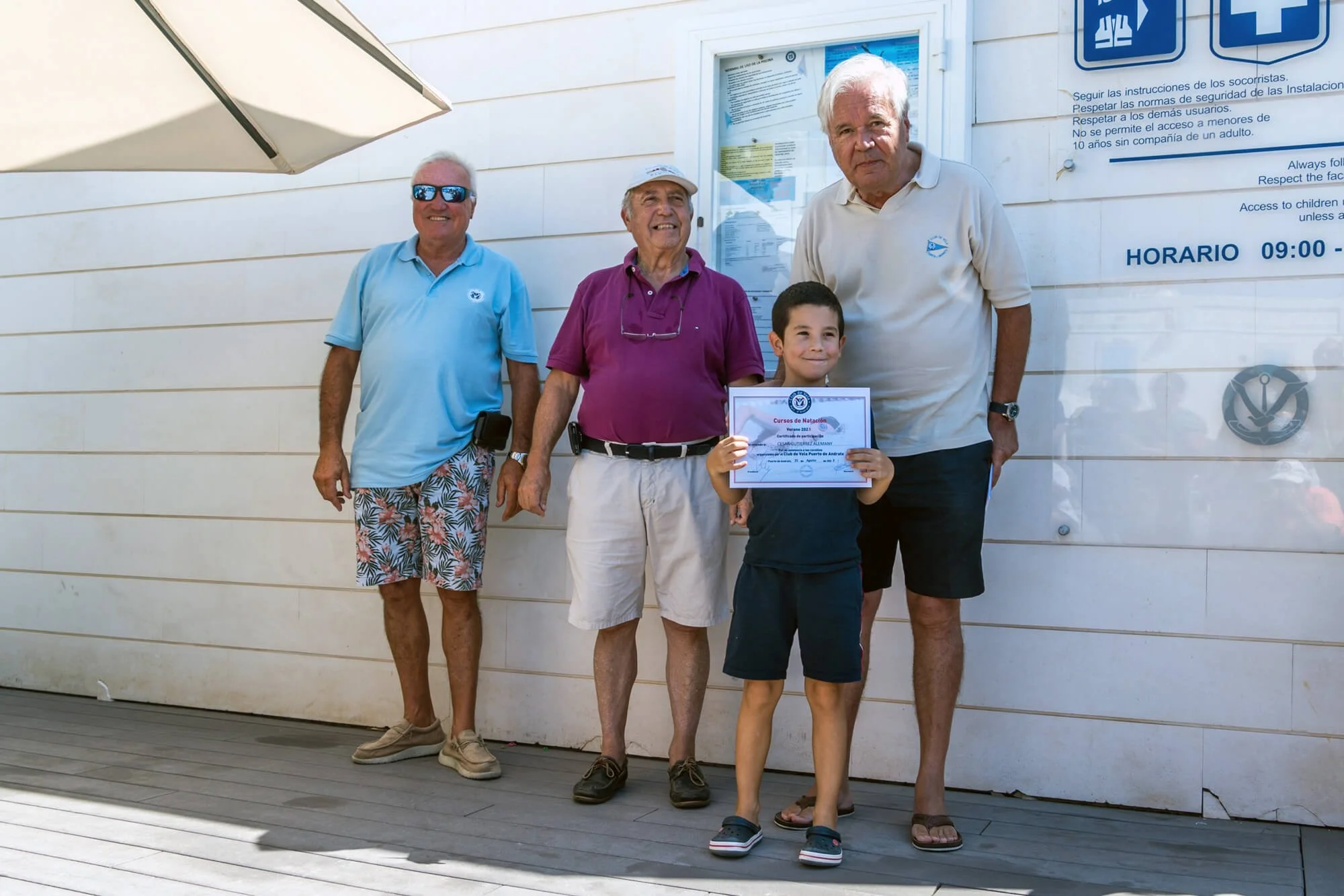 Grupo de cuatro personas, tres adultos mayores y un niño, sosteniendo un certificado en una ceremonia de premiación en un lugar al aire libre con fondo de pared de color claro y señales de instrucciones. Todos sonrientes, vistiendo ropa casual y de v
