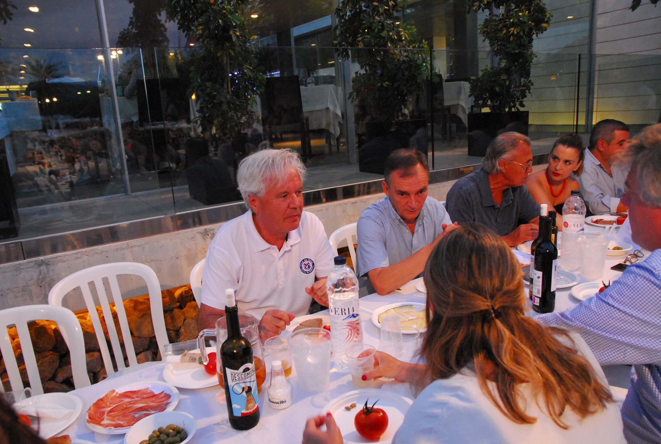 Grupo de personas cenando en una mesa con comida y bebidas en un ambiente interior con ventanas y plantas verdes.