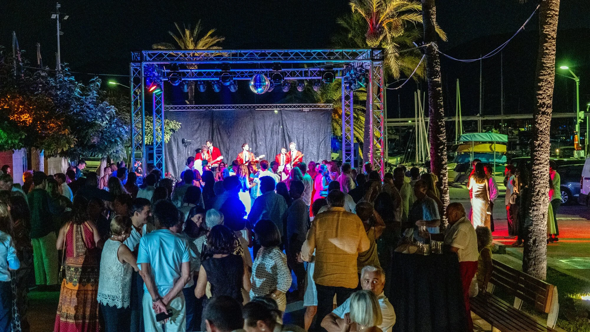 Concierto en la noche con público en un evento al aire libre, músicos vestidos con trajes rojos tocando en un escenario iluminado, palmeras y barcos en un muelle de fondo.