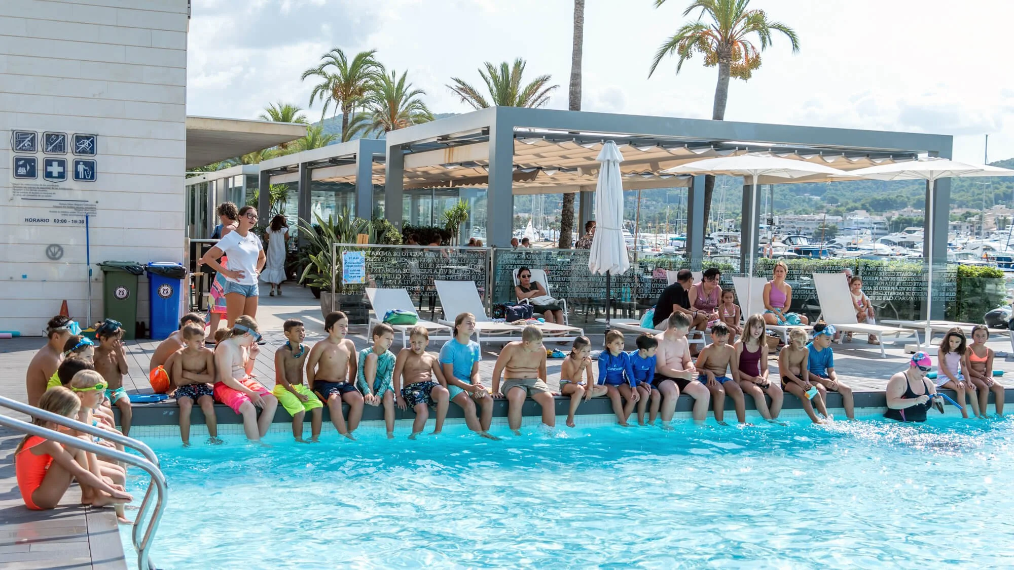 Niños sentados junto a la piscina después de nadar, algunos con gafas y gorros de natación, en un día soleado con palmeras y un comedor en el fondo.