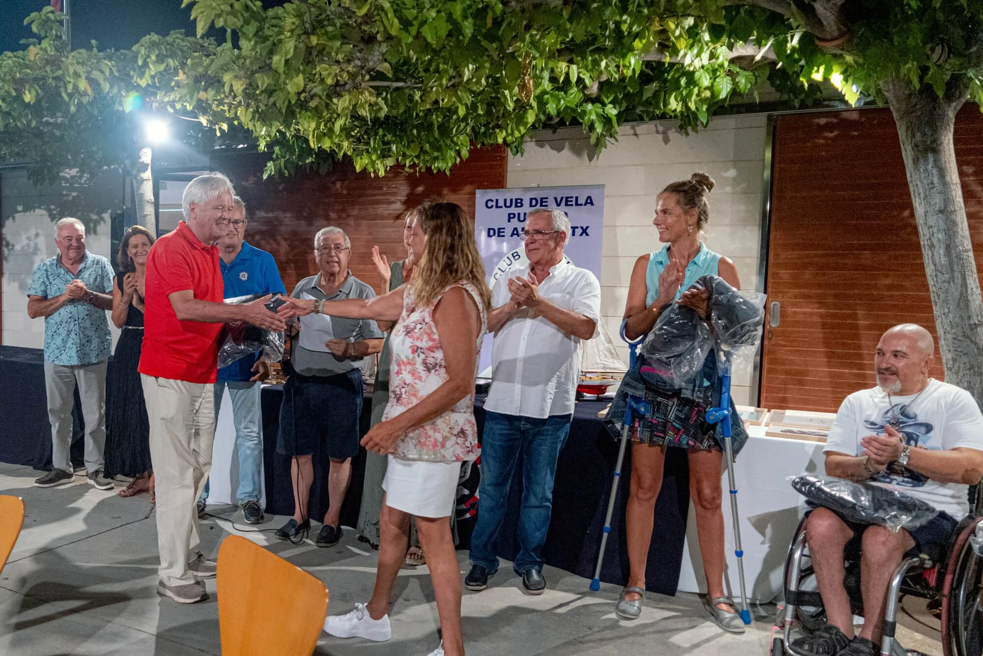 Grupo de personas en una ceremonia al aire libre, entregando un premio a una mujer con cabellera rizada, en una reunión social nocturna junto a árboles y una estructura de madera y metal.
