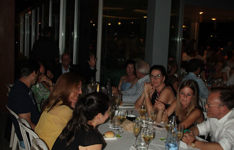 Grupo de personas en una comida en un restaurante, algunas sonrientes y brindando, en un ambiente animado y social.