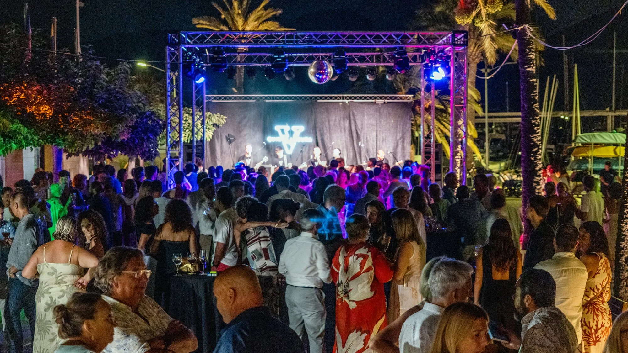 Escenario con banda tocando en concierto en una noche con muchas personas disfrutando la música, rodeado de palmeras y árboles