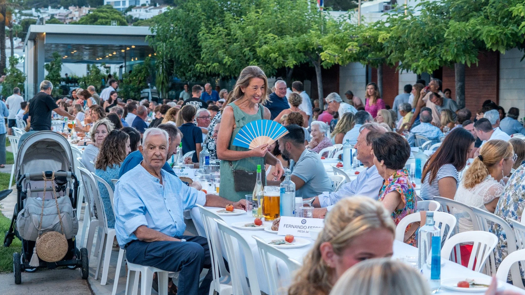 Reunión social al aire libre con muchas personas sentadas en mesas, sirviéndose comida y bebidas, en una celebración por la tarde, con árboles y un edificio en el fondo.