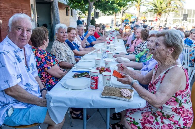 Grupo de personas mayores sentadas en una mesa larga al aire libre, disfrutando de una comida o reunión social.