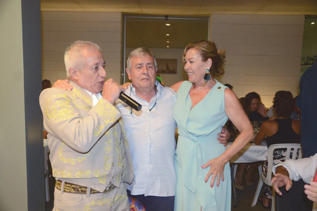 Tres personas en un evento social, una de ellas cantando con micrófono y las otras dos sonriendo, en un ambiente cerrado con mesas y otras personas al fondo.