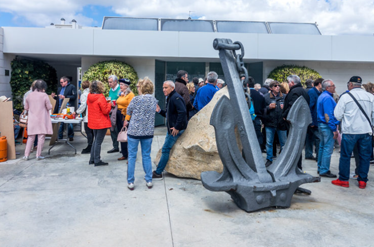 Grupo de personas en un evento al aire libre con una escultura de ancla y una roca delante de un edificio moderno.