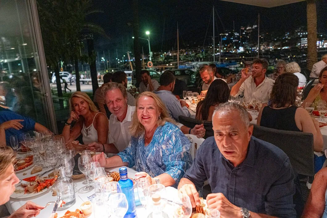 Grupo de personas cenando en un restaurante por la noche, con vista a un puerto con barcos y luces en el fondo.