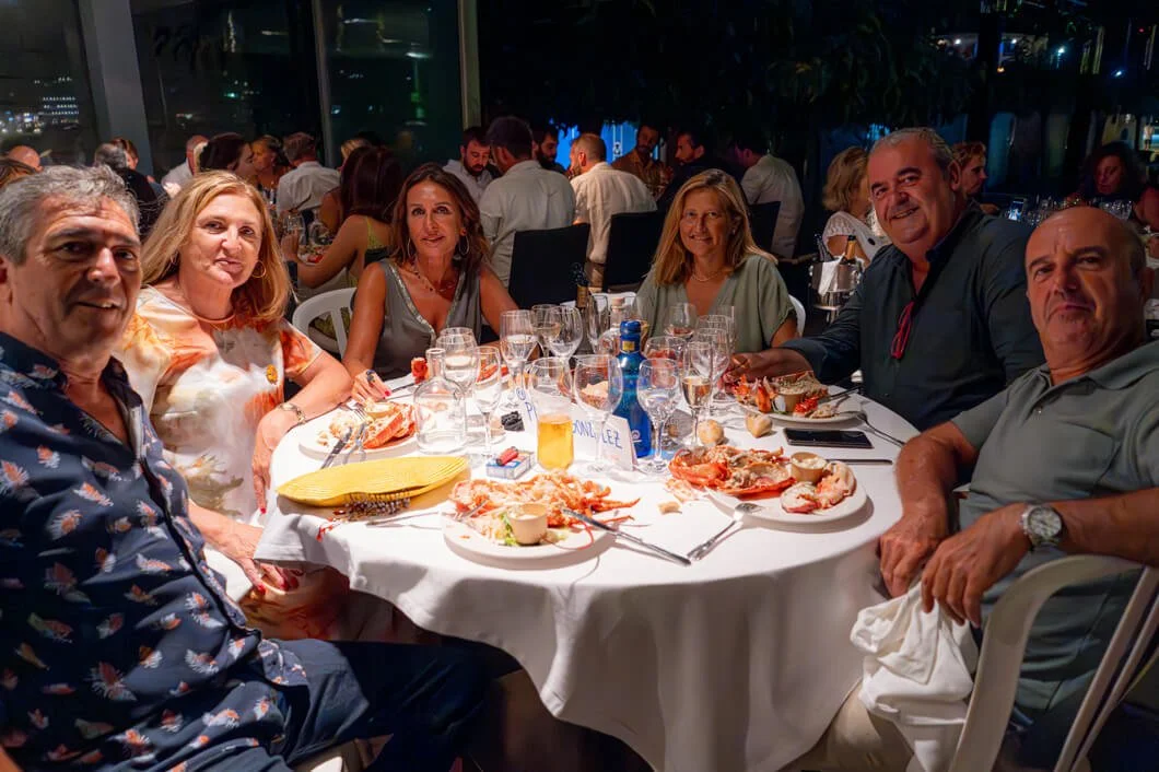 Grupo de personas sentadas en una mesa de banquete en un restaurante, disfrutando de comida y bebidas con muchos comensales en el fondo.