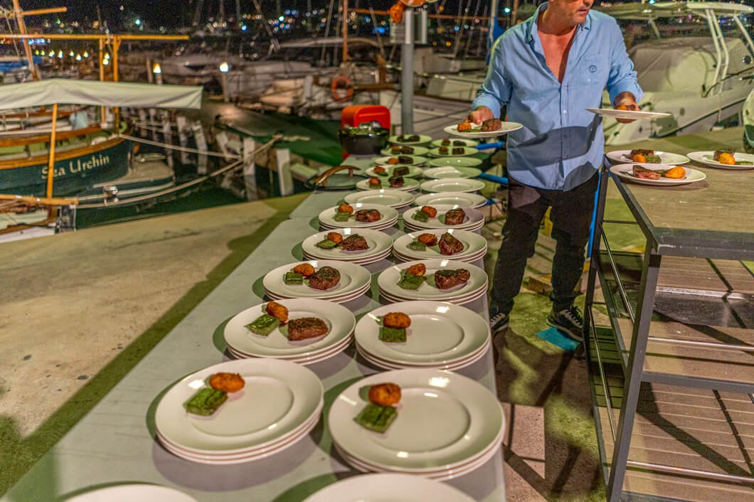 Persona sirviendo platos con carne, vegetales y aperitivos en una mesa al aire libre, en un muelle con barcos de fondo en un puerto por la noche.