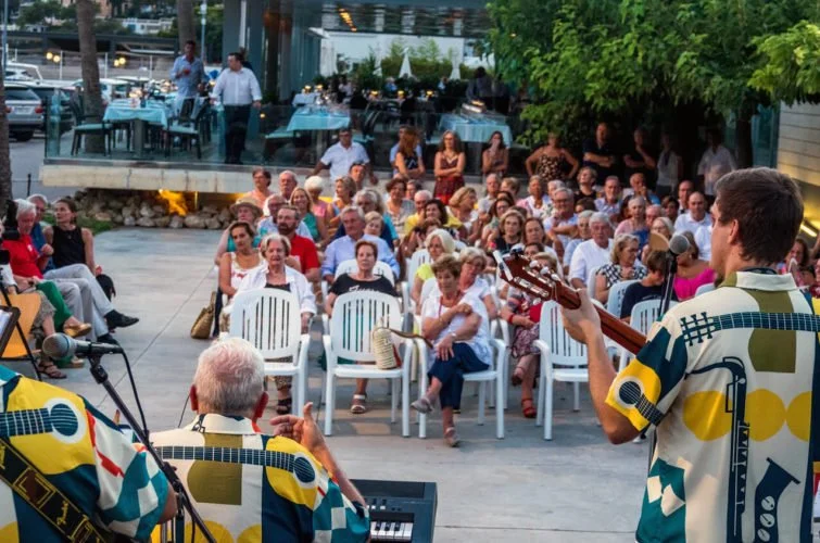 Un grupo de personas disfrutando de un concierto al aire libre con músicos tocando guitarras y teclado, en un espacio con mesas y sillas, con vegetación y un ambiente festivo.