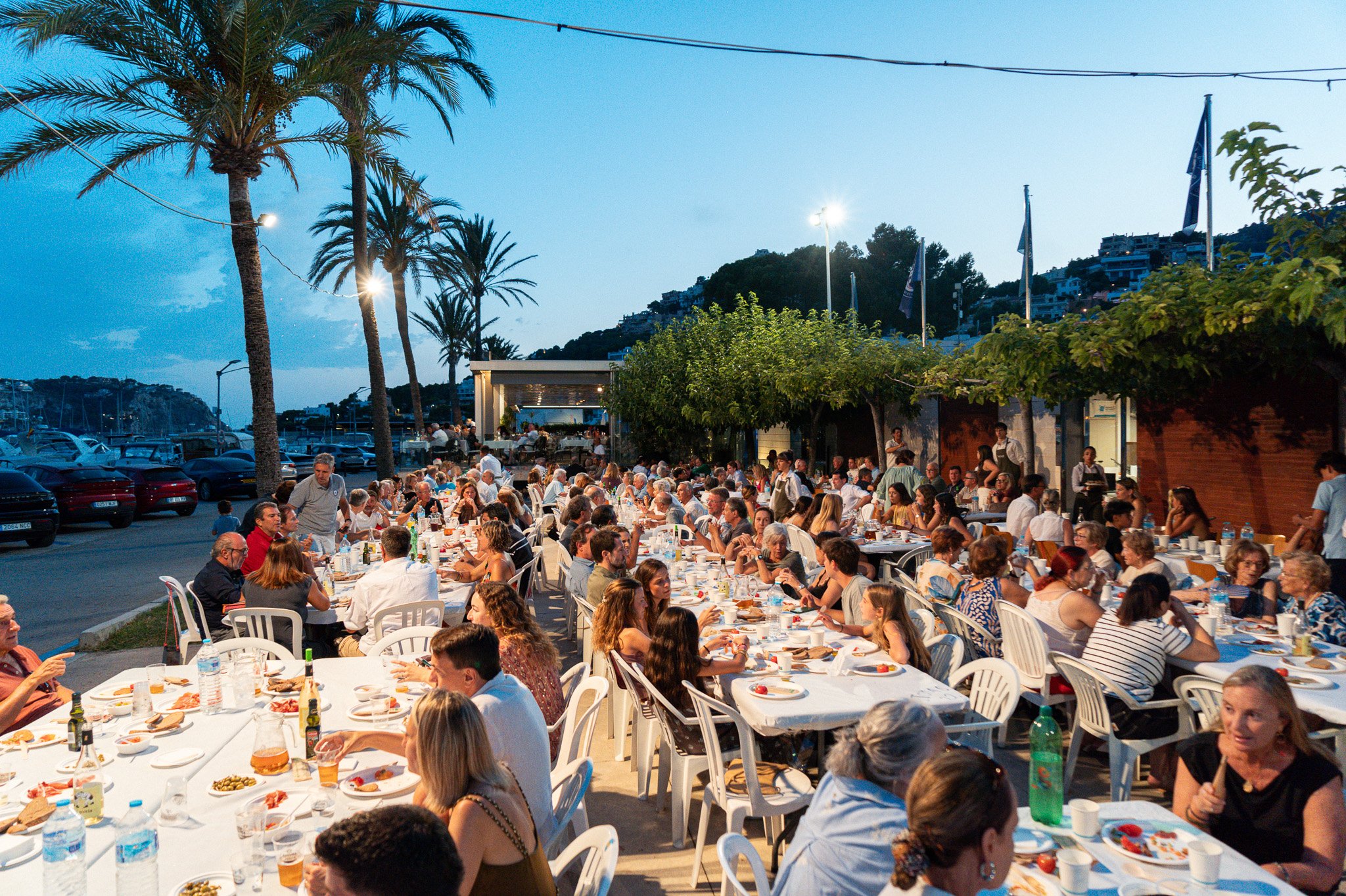 Una multitud de personas cenando en una terraza junto a un muelle con palmeras y montañas en el fondo, en la tarde o noche, en un ambiente festivo y social.