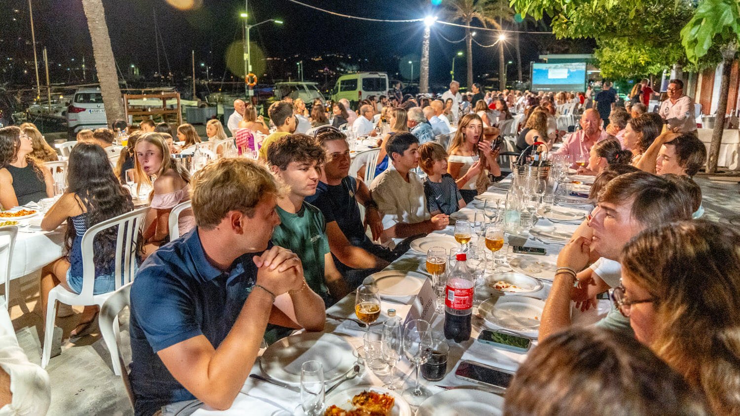 Una mesa larga llena de personas cenando en un evento al aire libre por la noche, con muchas personas participando y luz artificial iluminando el lugar.