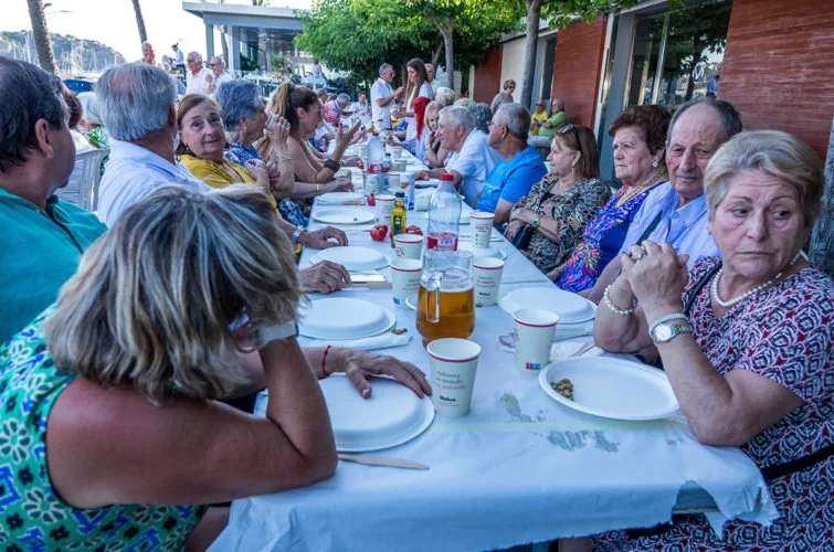 Grupo de personas mayores sentadas en una mesa larga al aire libre para una comida, con vasos y platos, algunas personas conversando y otras mirando hacia el frente.