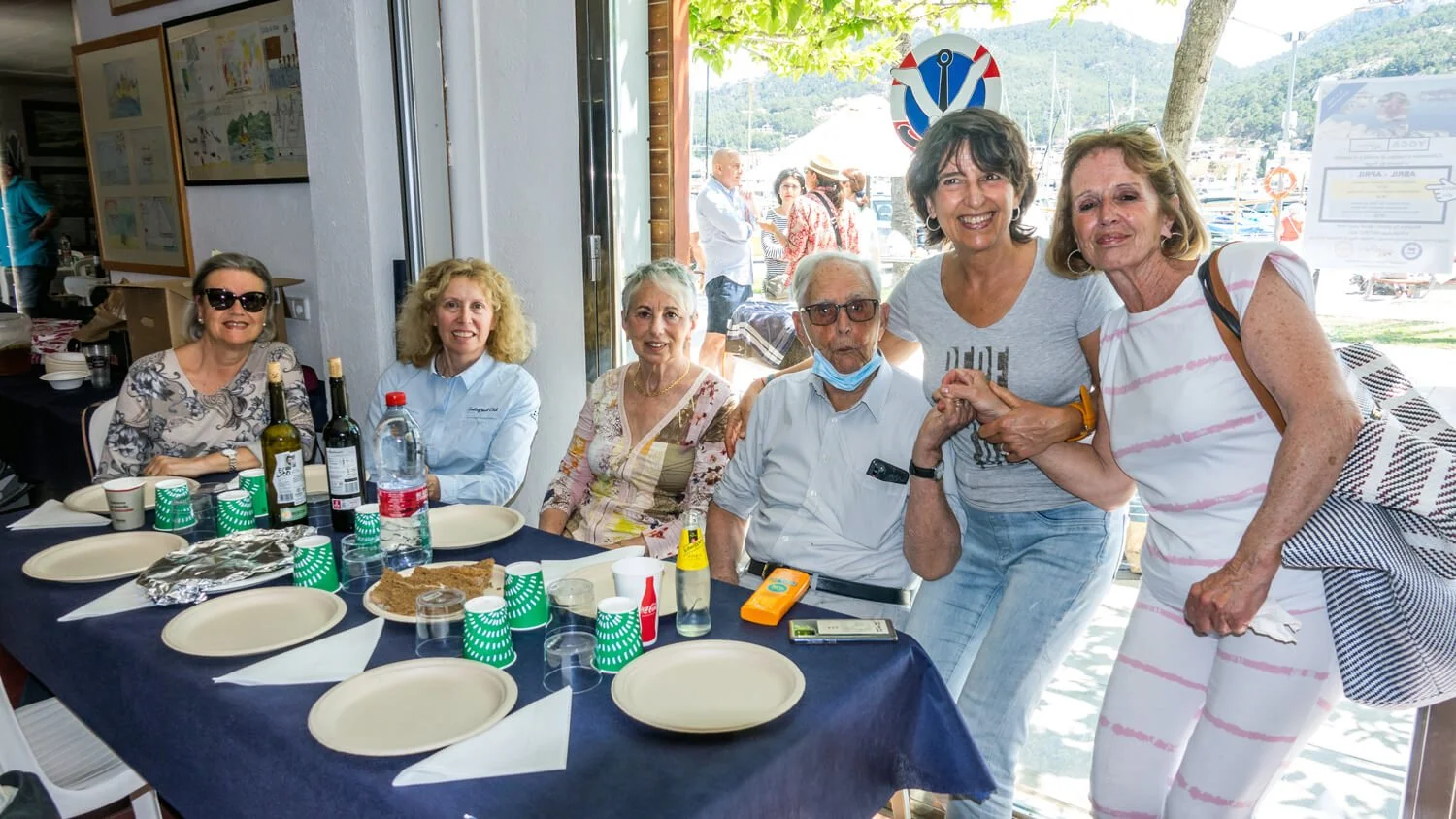 Grupo de personas en una comida al aire libre, algunos sentados y otros de pie, en un restaurante cerca del mar, con vista a barcos y montañas en el fondo.