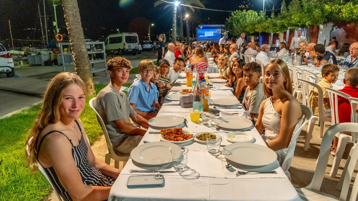 Reunión familiar o social en una comida al aire libre por la noche, con muchas personas sentadas en una larga mesa se ve comida, bebidas y un ambiente festivo.