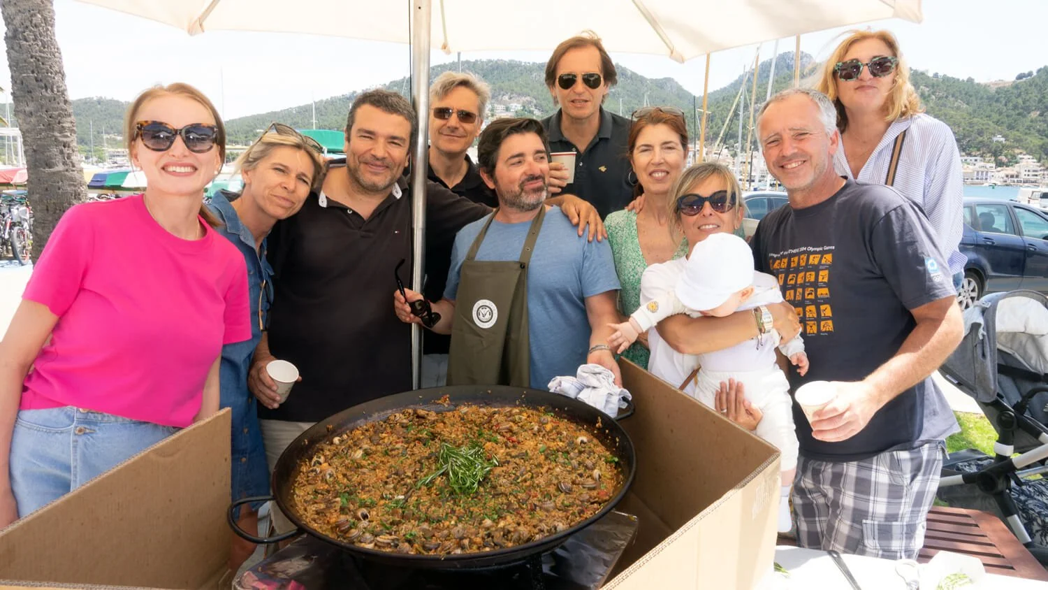 Grupo de personas en una reunión al aire libre con un plato grande de comida en el centro, en un entorno de puerto con montañas y barcos de fondo.