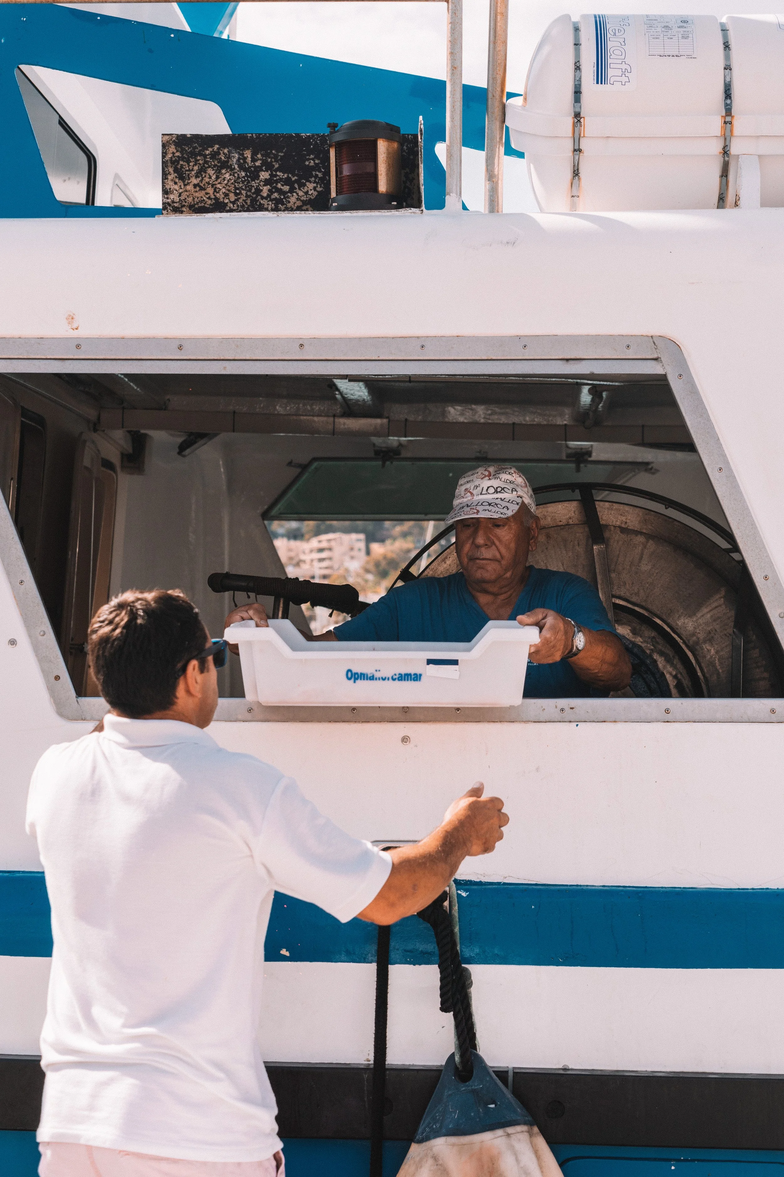 Un hombre con gorra azul atendiendo en una ventanilla de un barco, mientras otra persona en camisa blanca, vista de espaldas, le entrega un objeto con una cuerda. En la parte superior se ve parte del barco y un tanque.