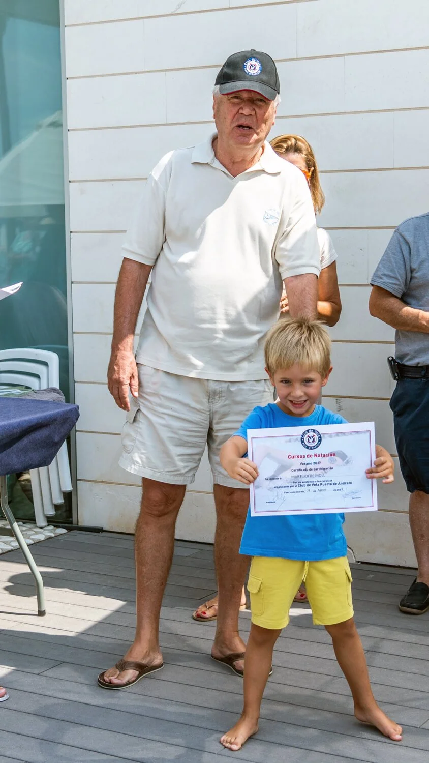 Un niño sonriente sostiene un certificado de participación en un curso de natación, con un adulto mayor con gorra y camiseta blanca a su lado, durante una ceremonia al aire libre.