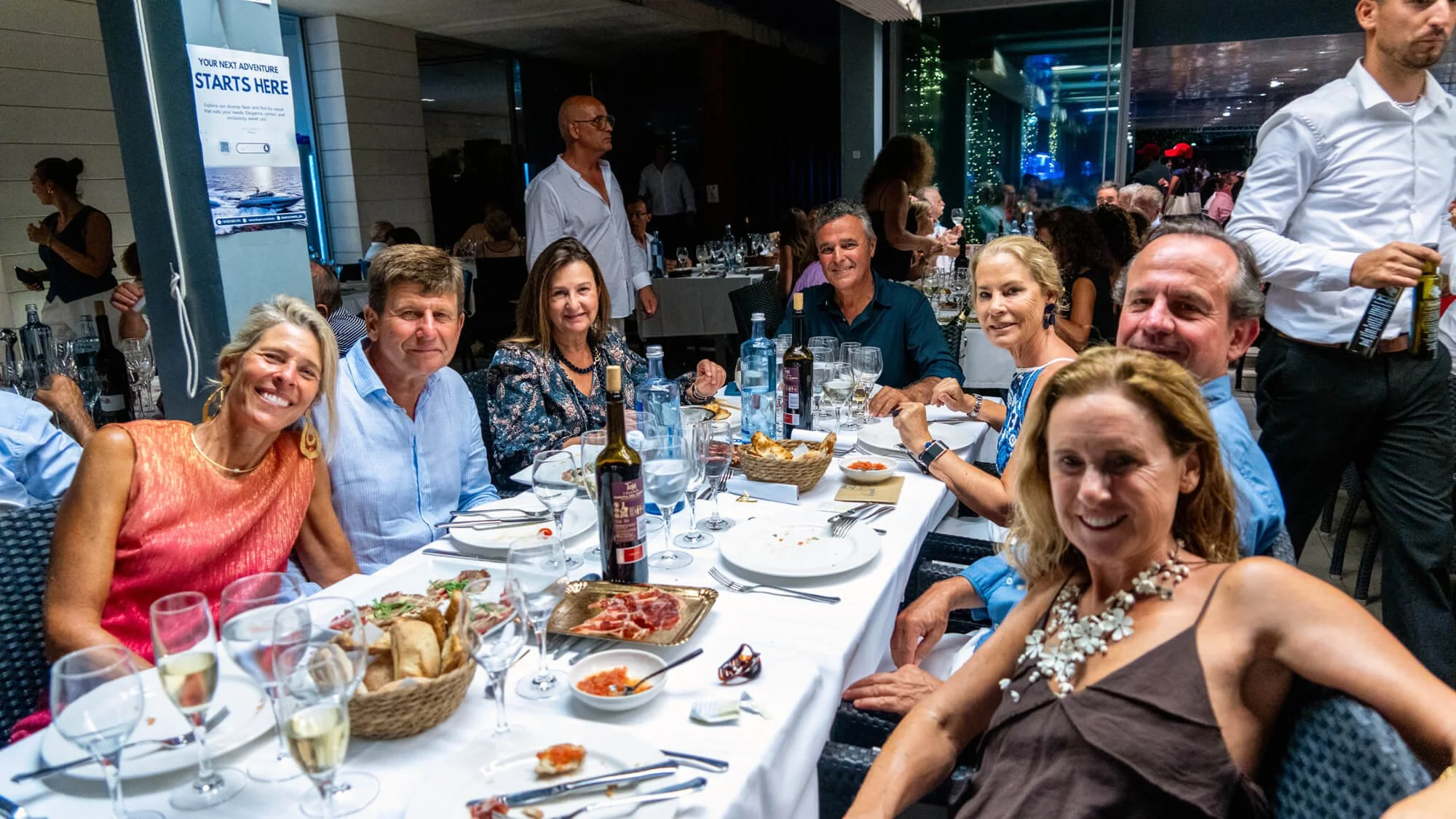 Grupo de personas disfrutando de una comida en un restaurante elegante, algunos sonriendo y otros serviendo su comida, con platos de comida y bebidas en la mesa.
