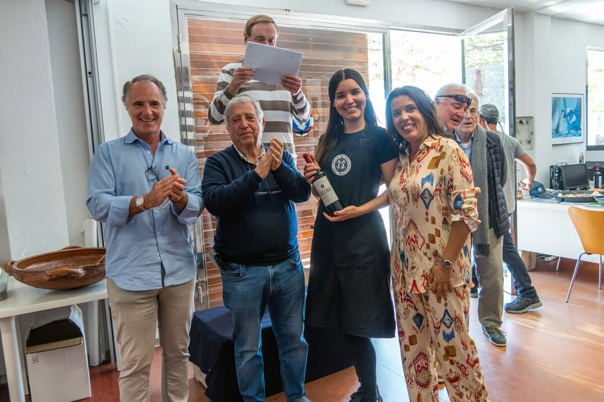 Grupo de personas en una ceremonia, la mujer en el centro recibe una botella de vino de una mujer con uniforme de evento, en un ambiente interior bien iluminado.